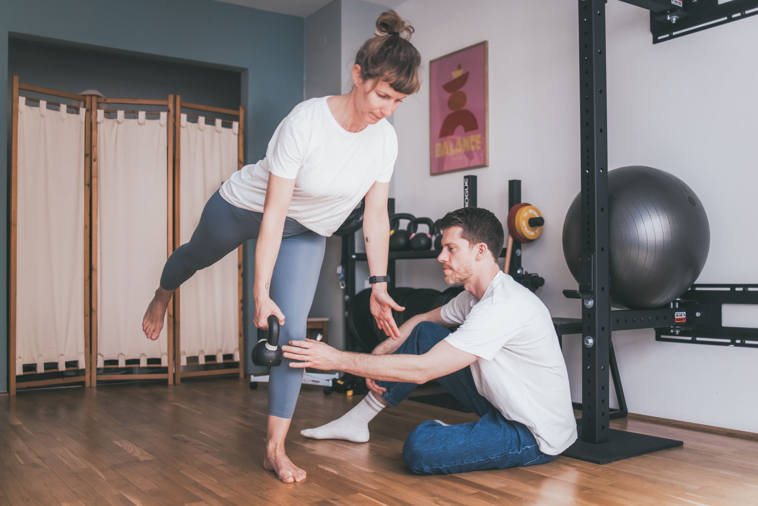 Patientin steht auf einem Bein mit einem Gewicht in der Hand und Daniel überprüft ihr Knie