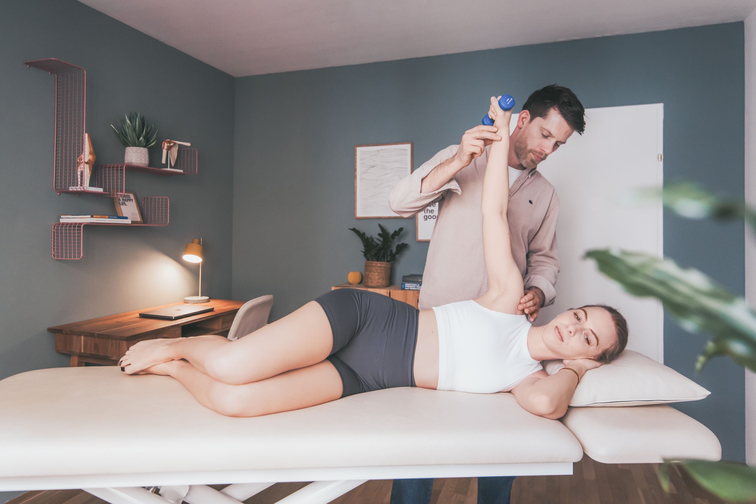 Daniel führt die Schulter einer Patientin, welche eine Übung auf der Therapieliege mit einer Hantel durchführt.