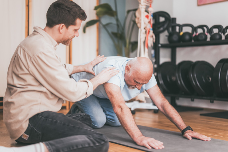 Daniel unterstützt Patienten bei einer Übung auf der Matte. Daniel unterstützt einen Patienten bei einer Übung.